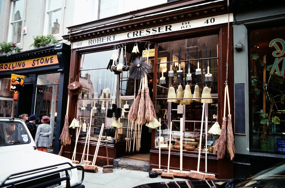 The storefront of Robert Cresser on Marylebone High Street displays a variety of cleaning tools and equipment arranged outside, including traditional straw and bristle brushes with long wooden handles, some with cloth or bristle heads hanging for display. The shop's large glass window showcases a well-maintained interior with warm lighting, highlighting cleaning or home decor items, while the exterior features a dark wood frame with clear signage. The surrounding street scene includes pedestrians and a parked car, with neighboring shop signs visible, reflecting a clean and inviting commercial retail environment consistent with professional cleaning standards promoted by Carpet Cleaning W1 for surface and deep cleaning of shop fronts and interiors.