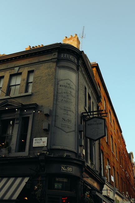 Exterior view of a historic building on Marylebone High Street in W1, with a rounded corner façade featuring large windows, decorative stonework, and signage for The Marylebonte. The building is constructed of brick and stone, painted in neutral tones, and illuminated by natural daylight. The street sign indicates the address as No. 98. Surrounding structures include other brick buildings with shopfronts and awnings. The scene reflects a busy retail area, emphasizing the importance of maintaining cleanliness and hygiene for commercial spaces, with Carpet Cleaning W1 offering surface cleaning and deep cleaning services to enhance the appearance and hygiene of shop interiors in Marylebone.