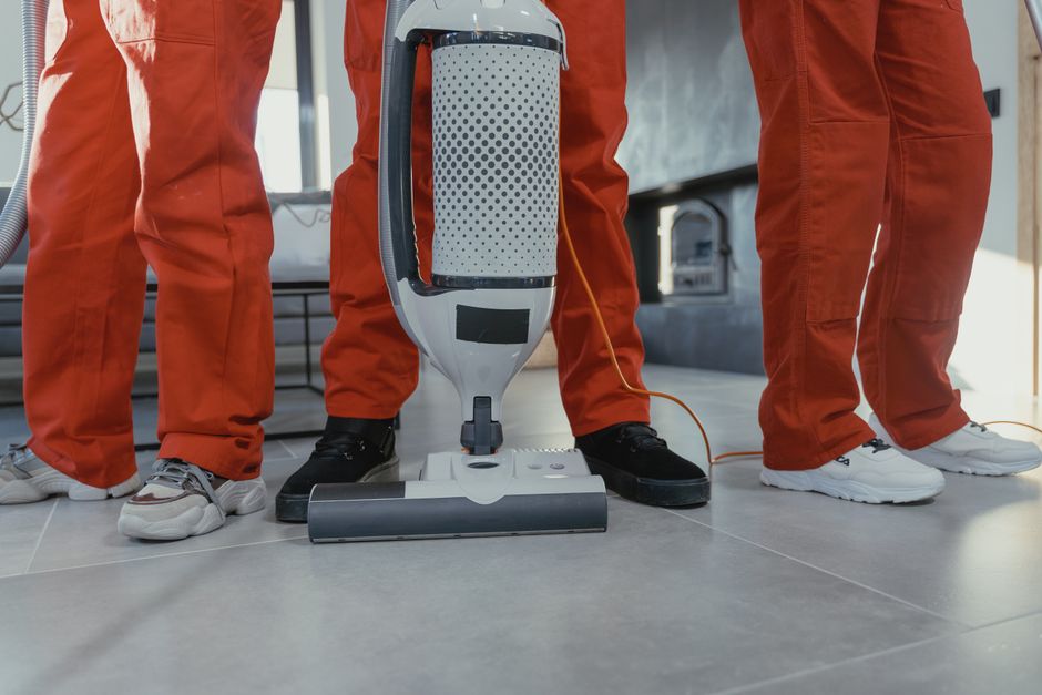 Three individuals dressed in orange protective suits and black shoes perform surface cleaning using a modern upright vacuum cleaner on a light gray tiled floor in a well-lit room, possibly a commercia