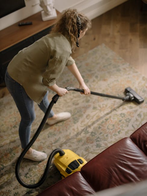 A person operating a yellow portable carpet cleaning or deep cleaning machine on a traditional patterned rug in a well-lit room. The focus is on the device's front grille and handle, with the user hol